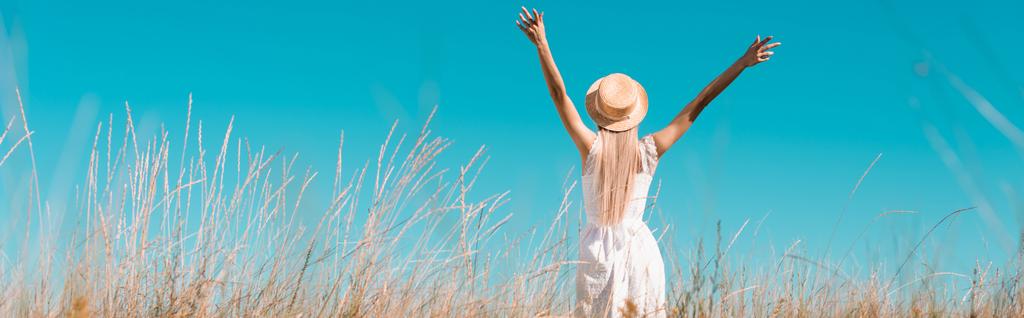 enfoque selectivo de la mujer rubia en vestido blanco y sombrero de paja de pie con las manos levantadas contra el cielo azul, imagen horizontal - Foto, imagen
