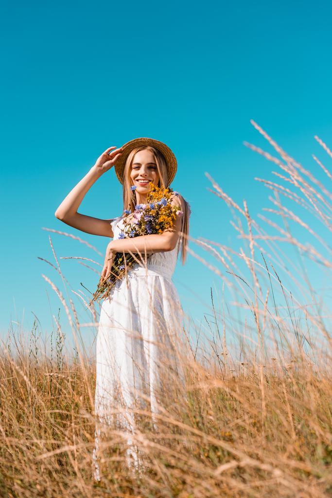 selective focus of young blonde woman in white dress holding wildflowers and touching straw hat while looking at camera - Photo, Image