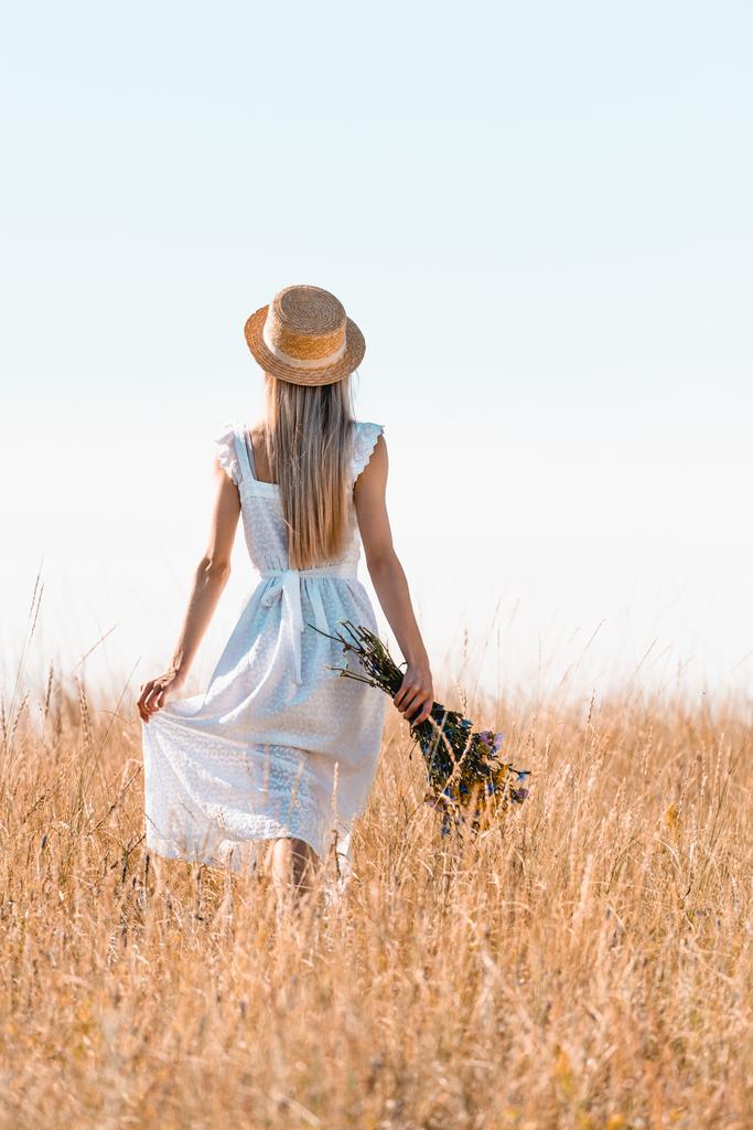 widok z tyłu kobieta w słomkowym kapeluszu dotykając białej sukienki podczas spaceru na trawiastej łące z bukietem dzikich kwiatów - Zdjęcie, obraz