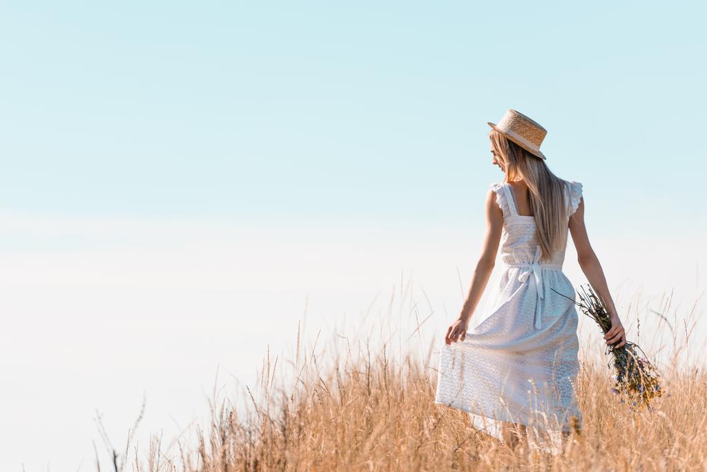 вид ззаду молодої жінки в солом'яному капелюсі, що торкається білого плаття, тримаючи дикі квіти на трав'янистому пагорбі
 - Фото, зображення
