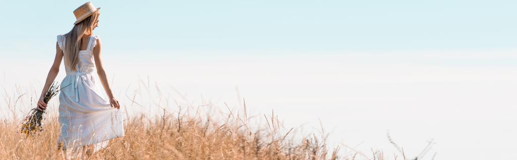 заголовок веб-сайту молодої жінки в білій сукні і солом'яному капелюсі, що стоїть на трав'янистому пагорбі з букетом диких квітів, вид ззаду
 - Фото, зображення