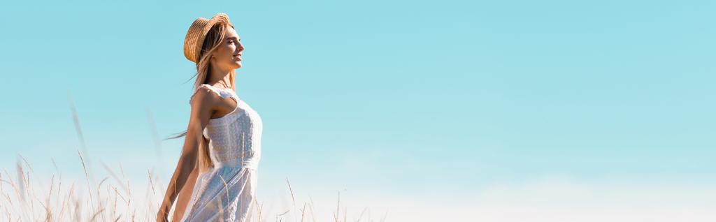 vista lateral de la mujer joven en vestido blanco y sombrero de paja de pie contra el cielo azul con las manos extendidas, tiro panorámico - Foto, imagen