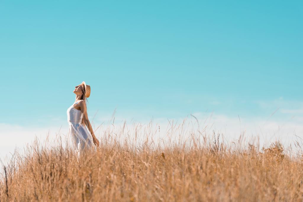 szelektív fókusz fiatal nő fehér ruha és szalma kalap álló kinyújtott kézzel és emelt fejjel ellen kék ég - Fotó, kép