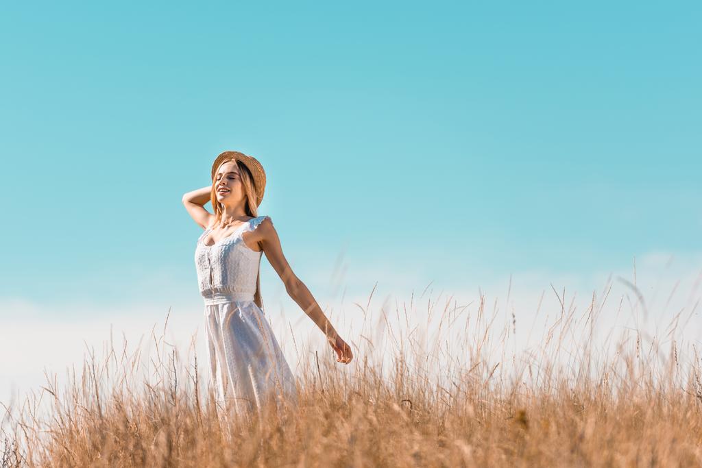 enfoque selectivo de la mujer sensual en vestido blanco tocando sombrero de paja mientras está de pie con las manos extendidas contra el cielo azul - Foto, Imagen