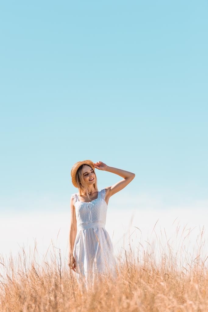 young woman in white dress standing on grassy hill, touching straw hat and looking away against blue sky - Photo, Image