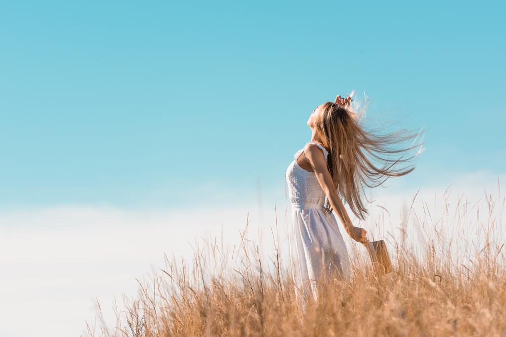 selective focus of blonde woman in white dress holding straw hat while standing with raised hand on grassy hill - Photo, Image