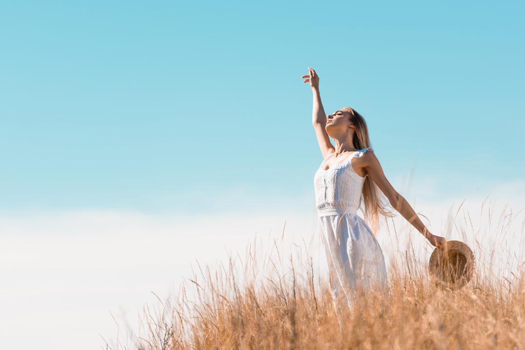 selectieve focus van blonde vrouw in witte jurk met strohoed terwijl staande met opgeheven hand op grasachtige heuvel - Foto, afbeelding