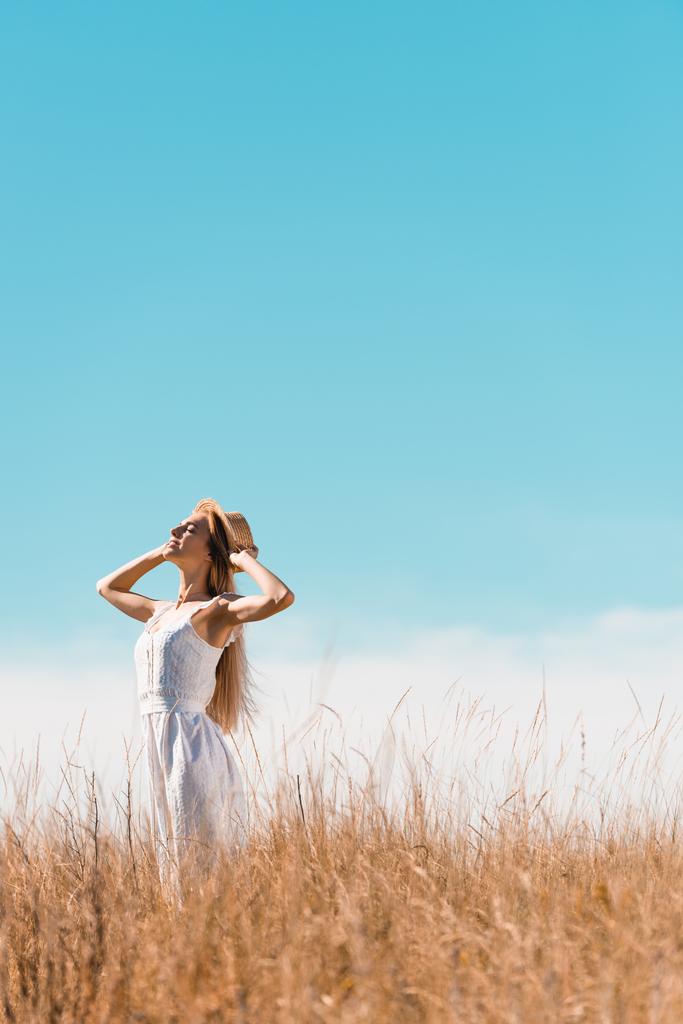 enfoque selectivo de la mujer con estilo en vestido blanco tocando sombrero de paja mientras está de pie con la cabeza levantada contra el cielo bullicio - Foto, imagen