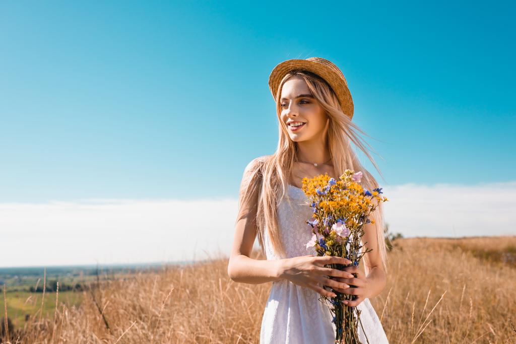 blonde vrouw in witte jurk en stro hoed met boeket van wilde bloemen terwijl weg te kijken tegen de blauwe lucht - Foto, afbeelding