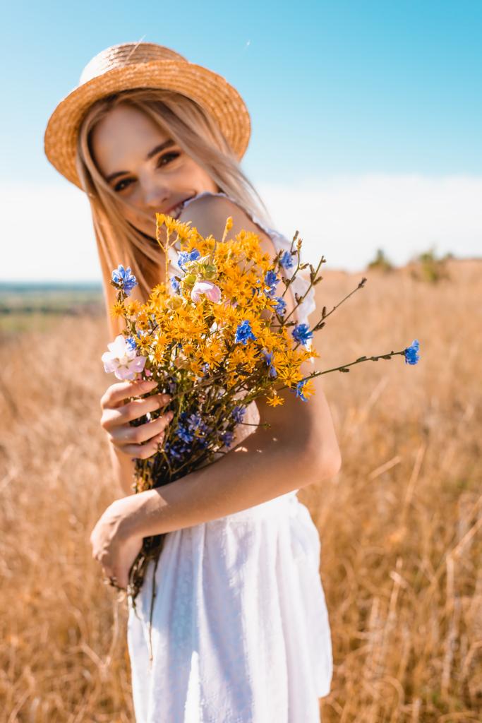 草むらの牧草地でカメラを見ながら野花を持つ麦藁帽子のブロンド女性の選択的な焦点 - 写真・画像