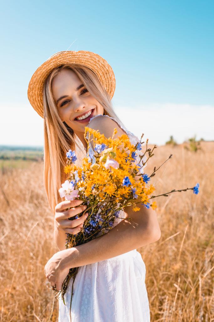 Çimenli tarlada kameraya bakarken elinde kır çiçekleri olan hasır şapkalı genç şık bir kadın. - Fotoğraf, Görsel
