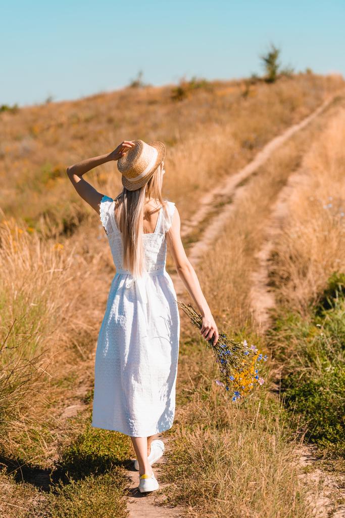 Вид сзади молодой женщины в белом платье, трогающей соломенную шляпу, идущей по дороге в поле с букетом полевых цветов - Фото, изображение