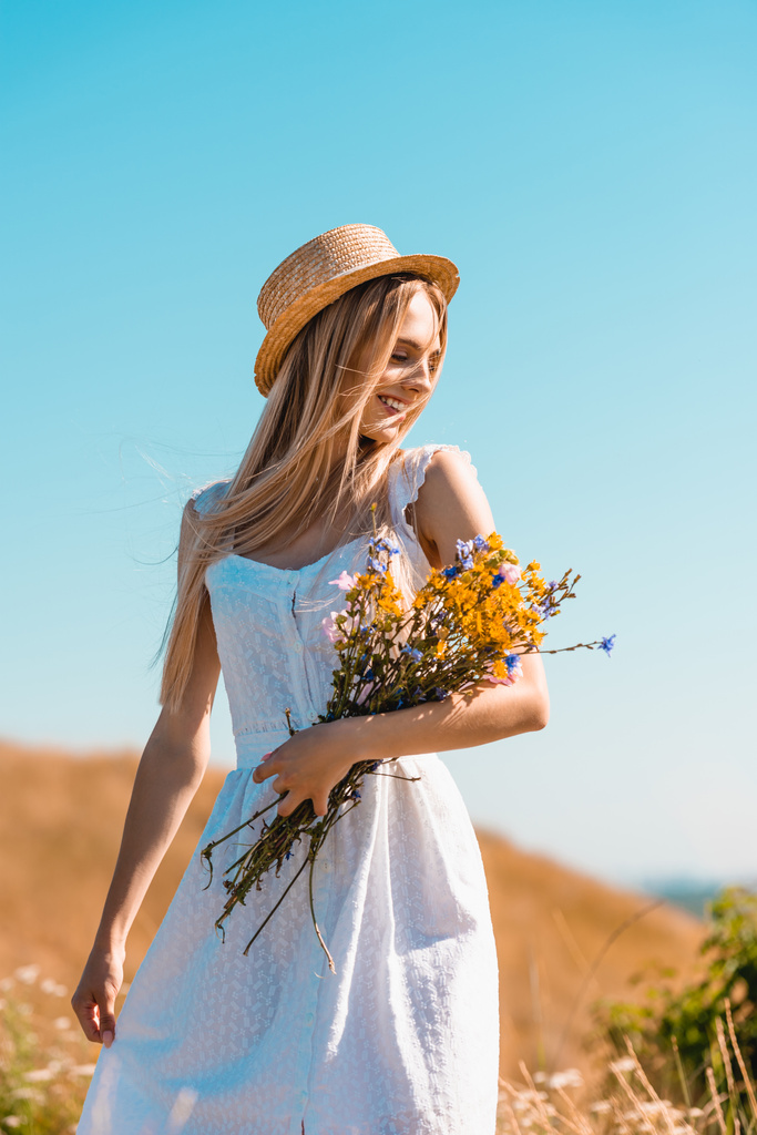 Beyaz elbiseli ve hasır şapkalı şehvetli kadın mavi gökyüzüne karşı kır çiçeği buketi tutuyor. - Fotoğraf, Görsel
