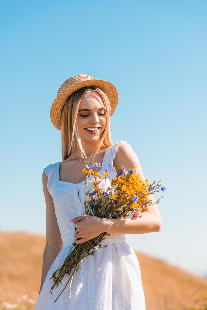 blonde woman in white dress and straw hat holding bouquet of wildflowers against blue sky - Photo, Image
