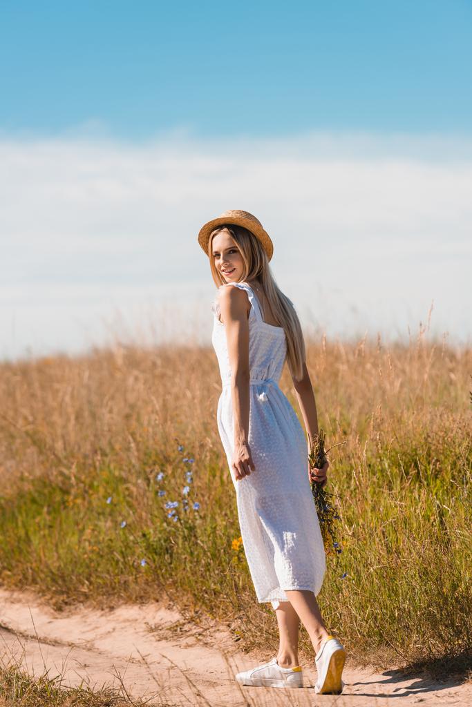 joven mujer rubia en sombrero de paja y vestido blanco sosteniendo flores silvestres y mirando a la cámara mientras camina por la carretera en el campo - Foto, Imagen