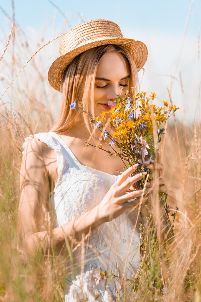 Hasır şapkalı şık bir kadının seçici odağı çimenlerdeki kır çiçeklerinin kokusunu alıyor. - Fotoğraf, Görsel