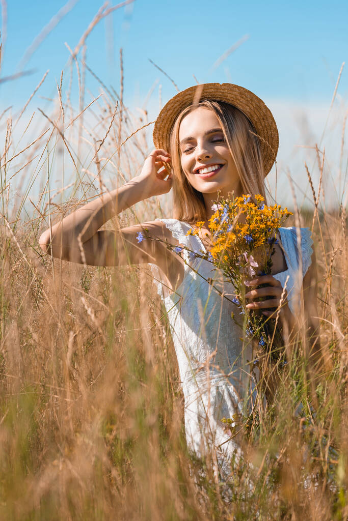 valikoiva painopiste nuori aistillinen nainen koskettaa olki hattu samalla tilalla luonnonvaraisia kukkia ruohoinen alalla - Valokuva, kuva