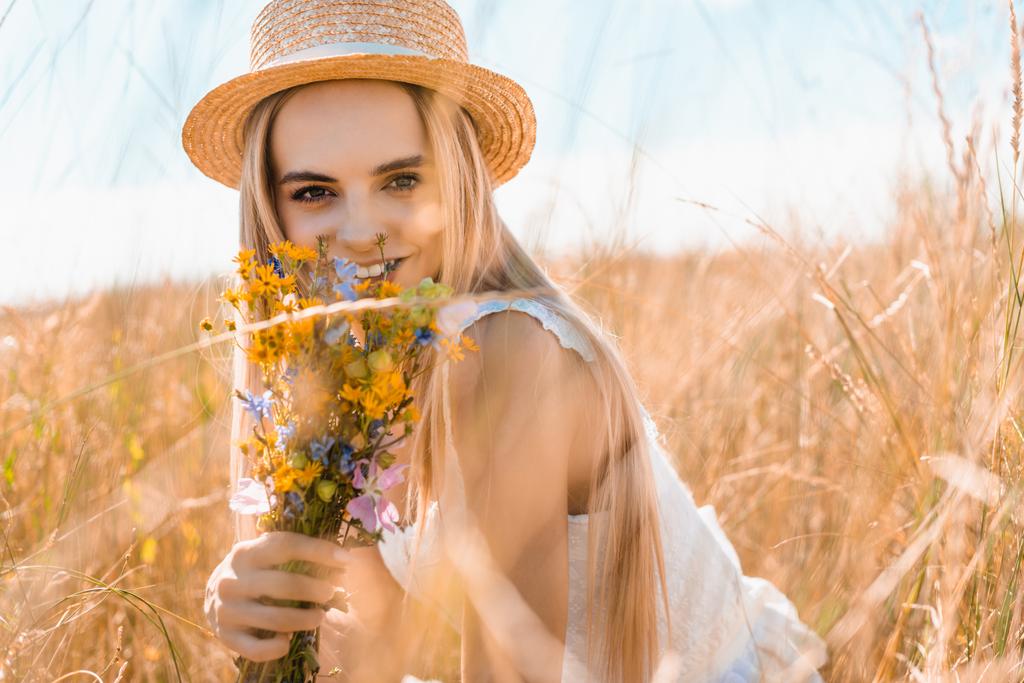 selektivní zaměření mladé ženy v slamáku drží divoké květiny a dívá se na fotoaparát na travnaté louce - Fotografie, Obrázek