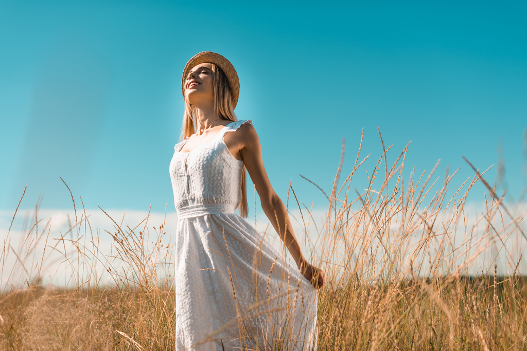 επιλεκτική εστίαση της νεαρής γυναίκας σε ψάθινο καπέλο αγγίζοντας λευκό φόρεμα, ενώ στέκεται με κλειστά μάτια σε χορταριασμένο λιβάδι - Φωτογραφία, εικόνα