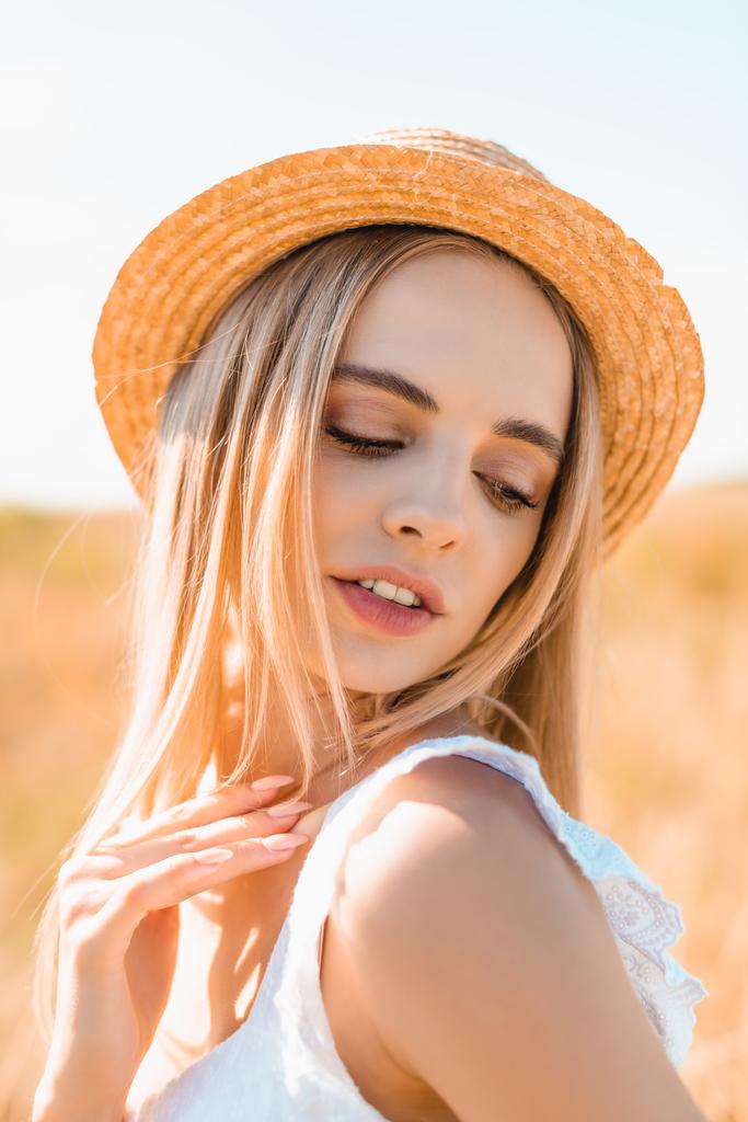 sensual blonde woman in straw hat and summer outfit touching shoulder in sunshine - Photo, Image