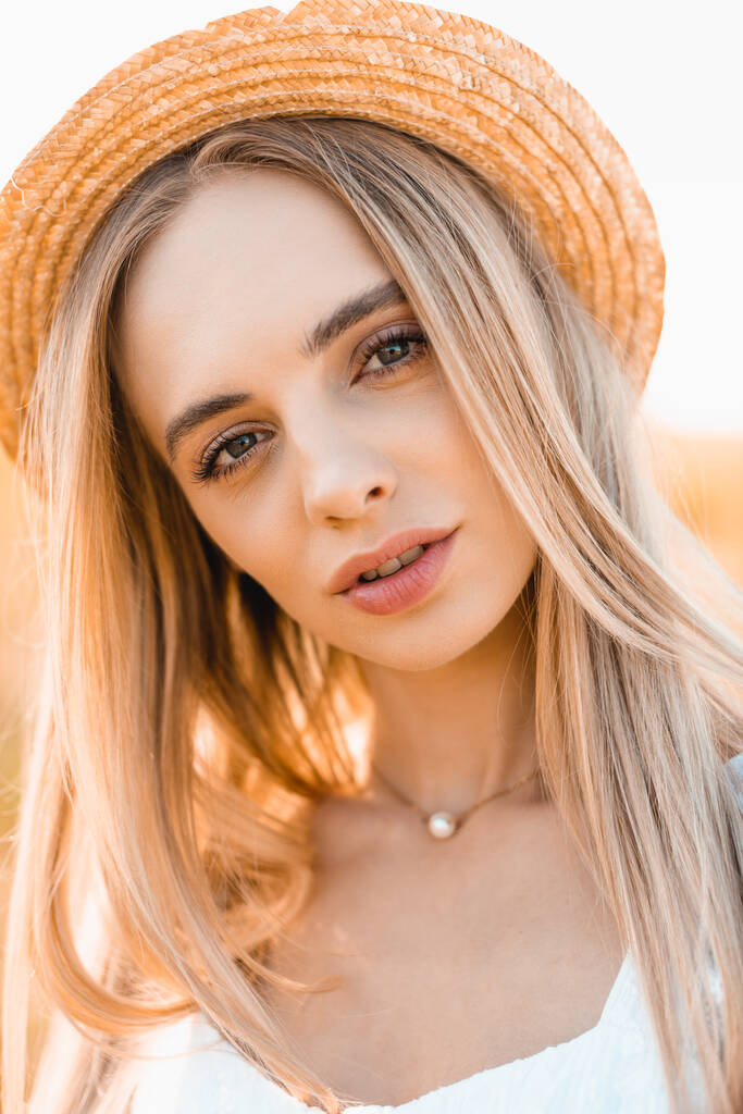 portret van jonge blonde vrouw in stro hoed kijken naar camera in zonneschijn - Foto, afbeelding
