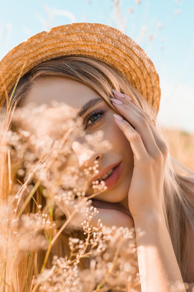 портрет мечтательной блондинки в соломенной шляпе, касающейся лица и смотрящей в камеру рядом с полевыми цветами - Фото, изображение