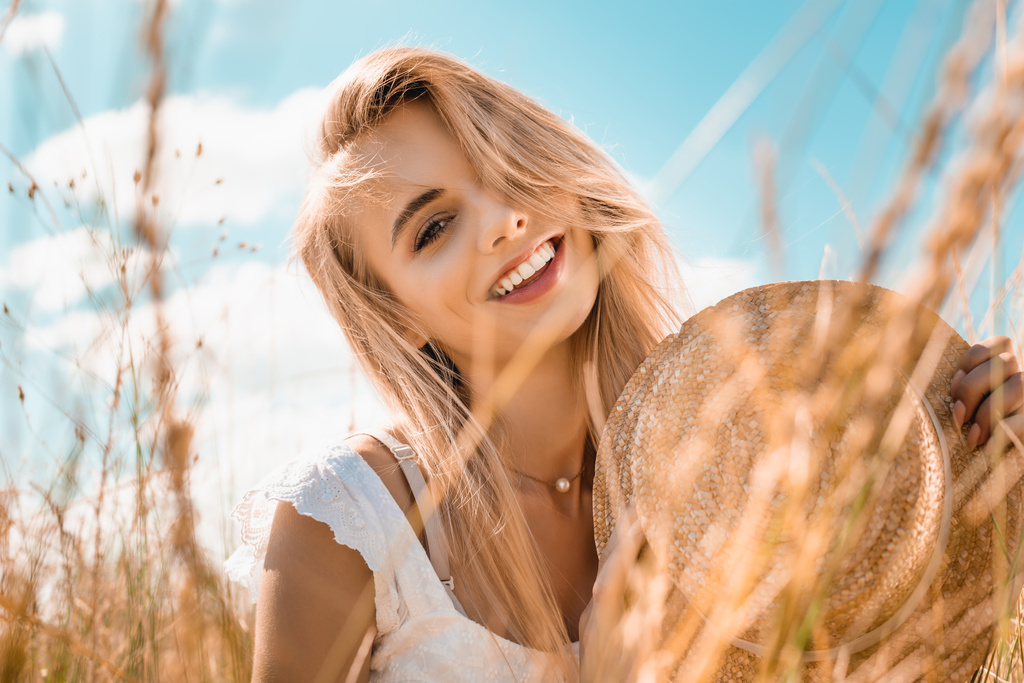 Selektiver Fokus der blonden Frau mit Strohhut und Blick in die Kamera im Gras vor blauem wolkenverhangenem Himmel - Foto, Bild
