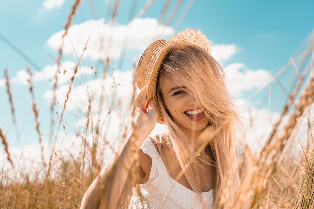 selektivní zaměření vzrušené blondýny žena dotýkající se slaměného klobouku a při pohledu na kameru proti modré oblačné obloze - Fotografie, Obrázek