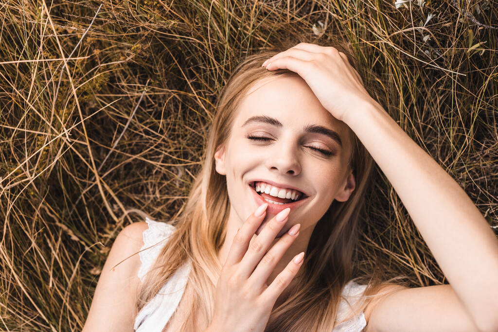 top view of excited blonde woman touching lips and forehead while lying on grass with closed eyes - Photo, Image