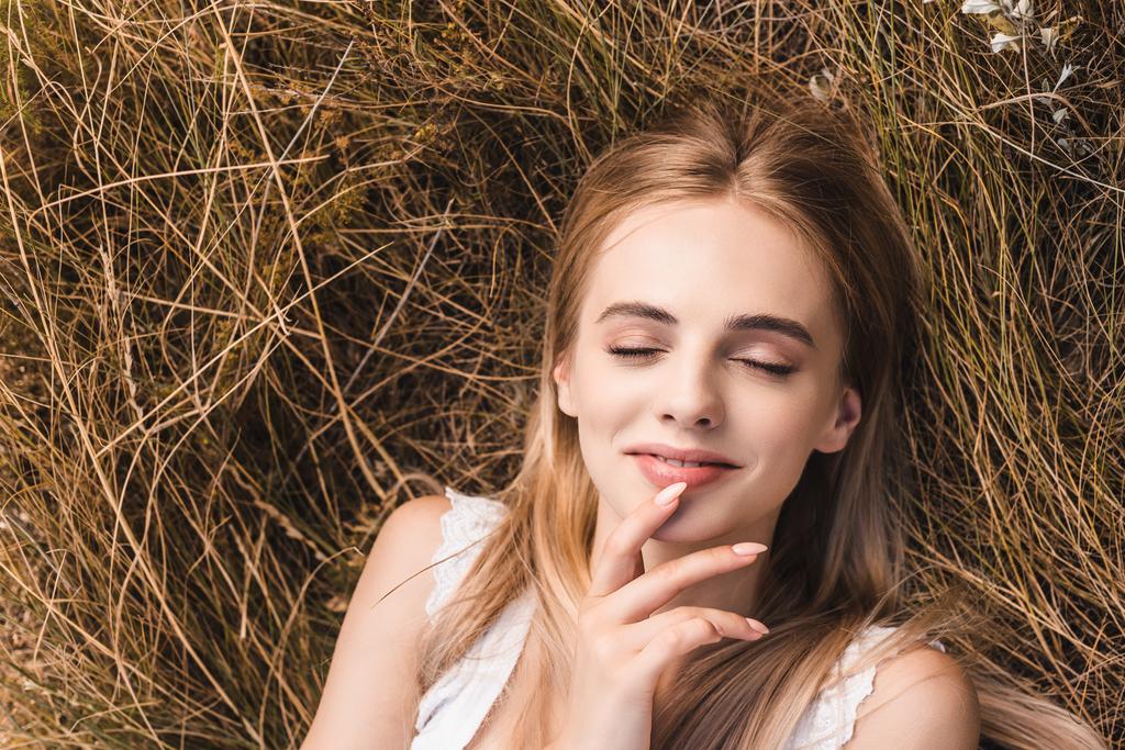 閉じた目で緑の芝生に横たわっている間に唇に触れる官能的なブロンドの女性のトップビュー - 写真・画像