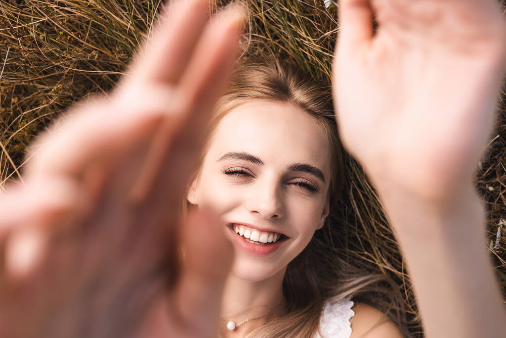 top view of excited blonde woman looking at camera while lying on grass with raised hands, selective focus - Photo, Image