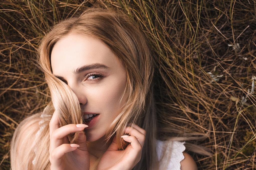 top view of sensual blonde woman lying on green grass, obscuring face with hair and looking at camera - Photo, Image