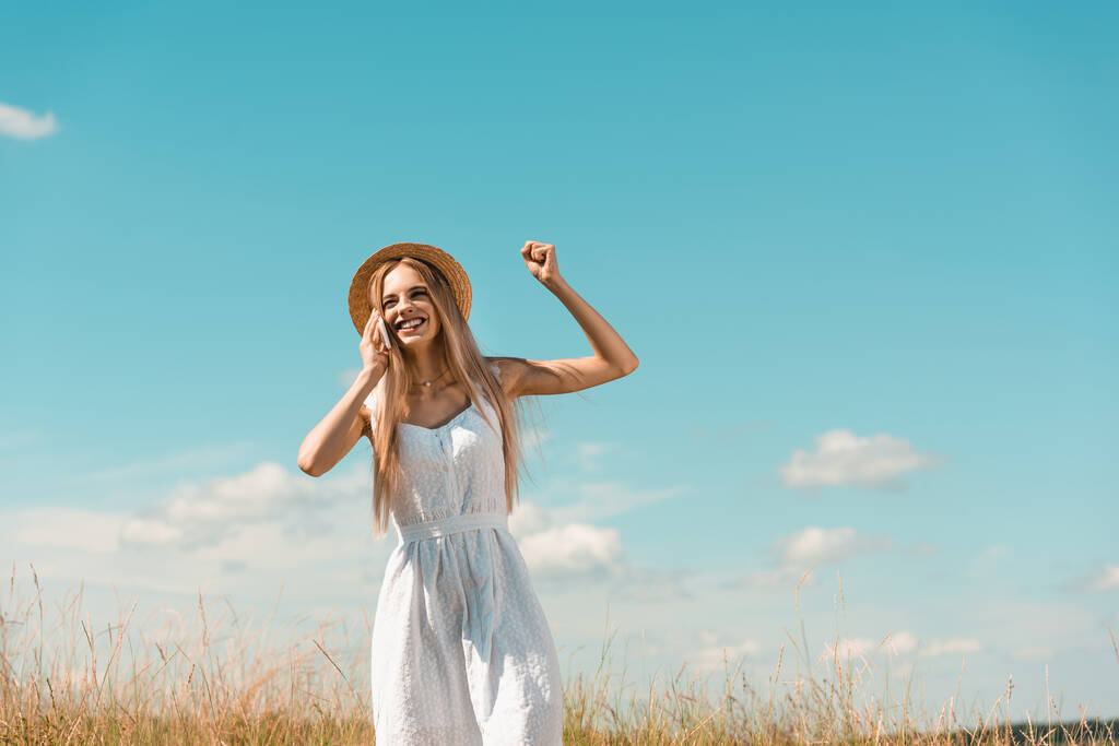 femme blonde excitée en robe blanche et chapeau de paille montrant geste gagnant tout en parlant sur smartphone contre le ciel bleu - Photo, image