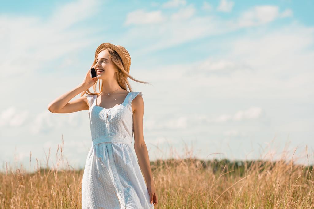 mujer joven en vestido blanco y sombrero de paja hablando en el teléfono inteligente mientras está de pie en el campo con los ojos cerrados - Foto, imagen