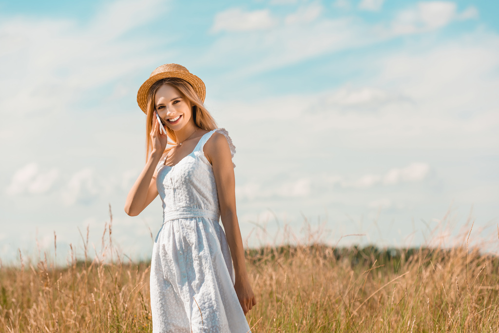 Beyaz elbiseli ve hasır şapkalı genç bir kadın mavi gökyüzüne karşı çayırda telefonla konuşurken kameraya bakıyor. - Fotoğraf, Görsel