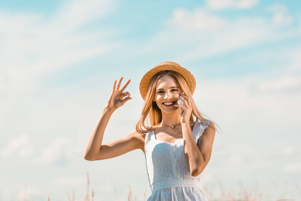 junge, aufgeregte Frau in weißem Kleid und Strohhut zeigt okay Geste, während sie mit dem Smartphone vor blauem Himmel spricht - Foto, Bild