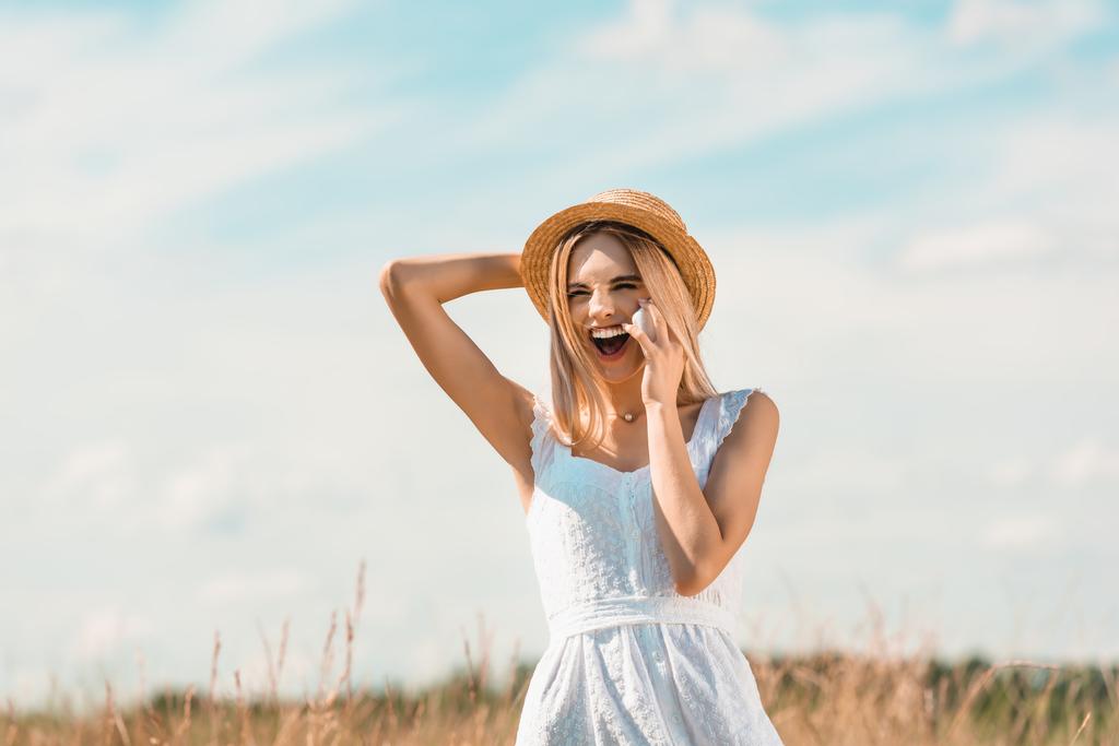 Beyaz elbiseli heyecanlı sarışın kadın hasır şapkaya dokunuyor ve mavi gökyüzüne gülüyor. - Fotoğraf, Görsel