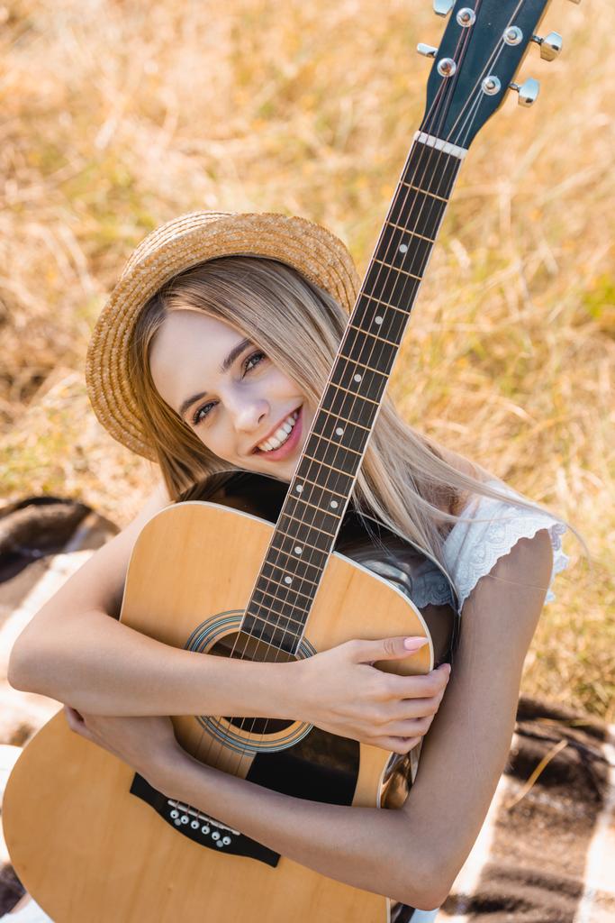 wysoki kąt widzenia blondynka kobieta w słomkowym kapeluszu siedzi na kocu w polu z gitarą akustyczną - Zdjęcie, obraz