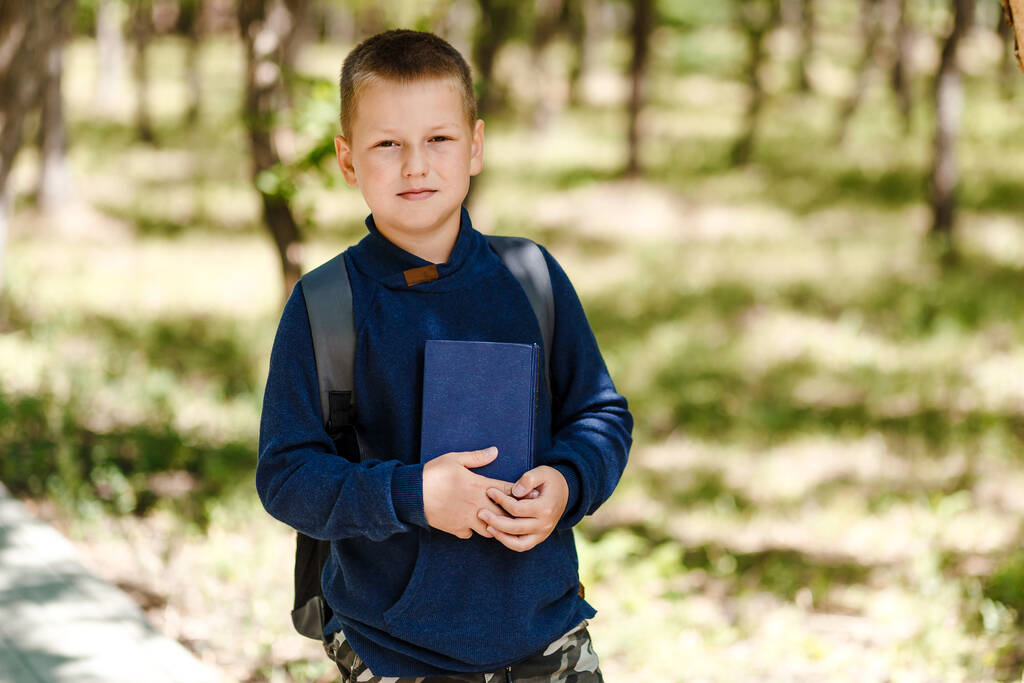 garçon de huit ans avec un sac à dos et un livre à la main à l'extérieur - Photo, image