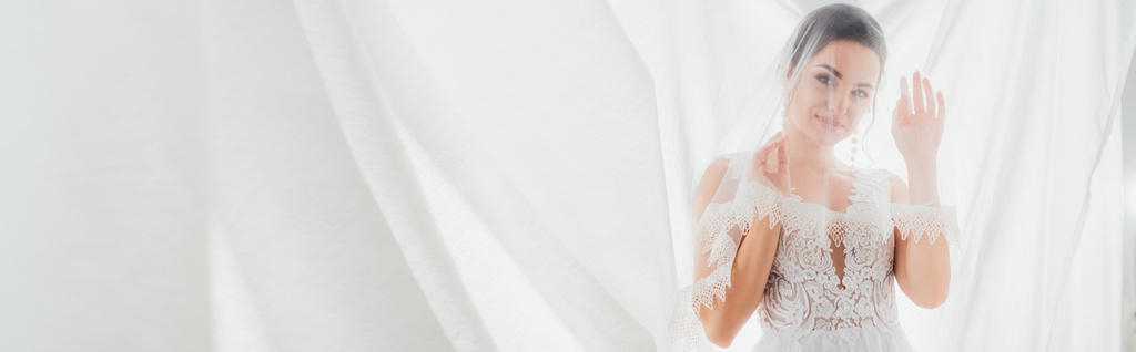 Horizontale oogst van bruid in trouwjurk en sluier kijken naar camera in de buurt van witte gordijnen  - Foto, afbeelding