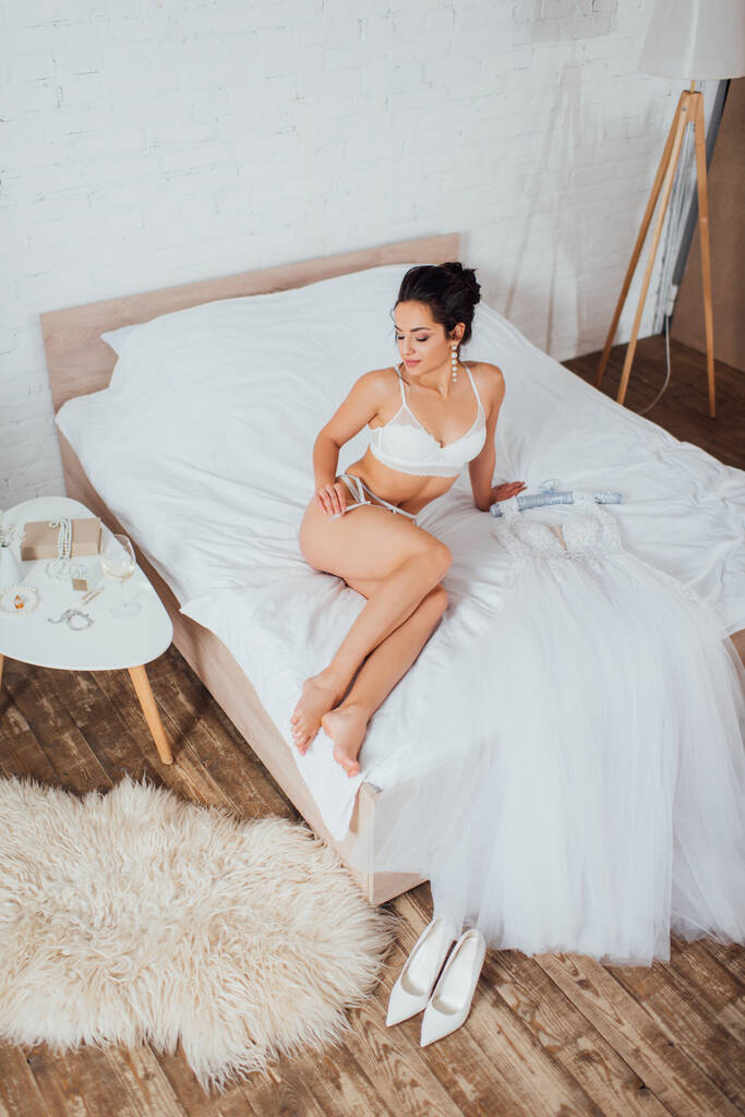 Sexy nevěsta ve spodním prádle dotýká podvazkový pás v blízkosti svatební šaty na posteli doma  - Fotografie, Obrázek