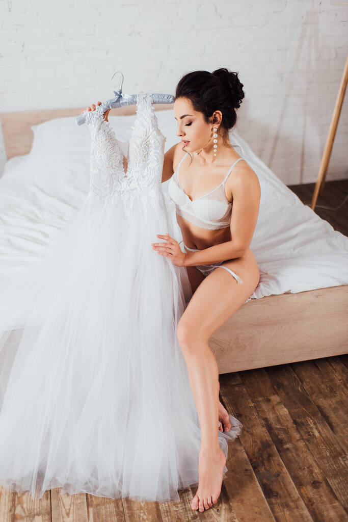 Morena novia en sujetador de encaje tocando vestido de novia en la percha en el dormitorio - Foto, Imagen