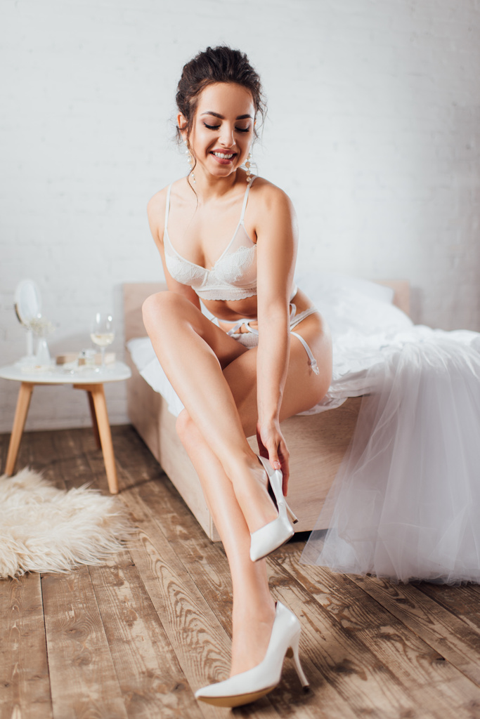 Selektivní zaměření nevěsty ve spodním prádle uvedení na boty v blízkosti svatební šaty na posteli  - Fotografie, Obrázek