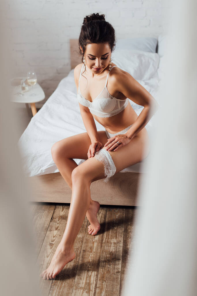 Foco seletivo de noiva sexy em lingerie e brincos de pérola colocando em liga de renda na cama
  - Foto, Imagem