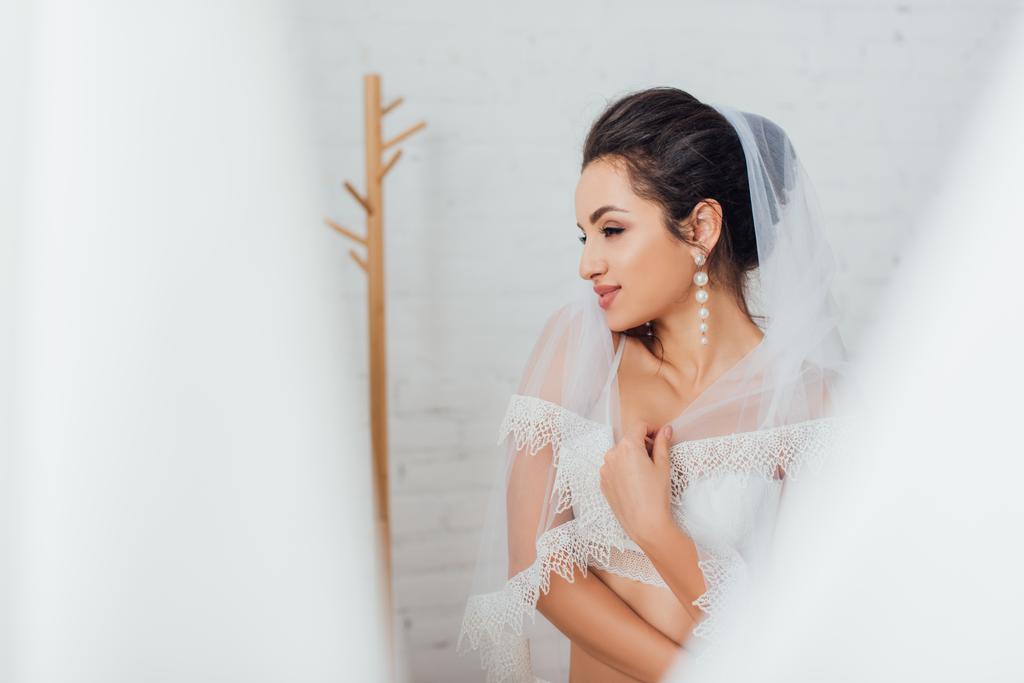 Выборочный фокус брюнетки невесты в бюстгальтере и жемчужной серьге носить кружевную вуаль и смотреть в сторону дома  - Фото, изображение