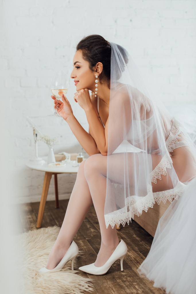 Foco seletivo da noiva em roupas íntimas e véu segurando vidro de vinho perto do vestido de noiva na cama
  - Foto, Imagem