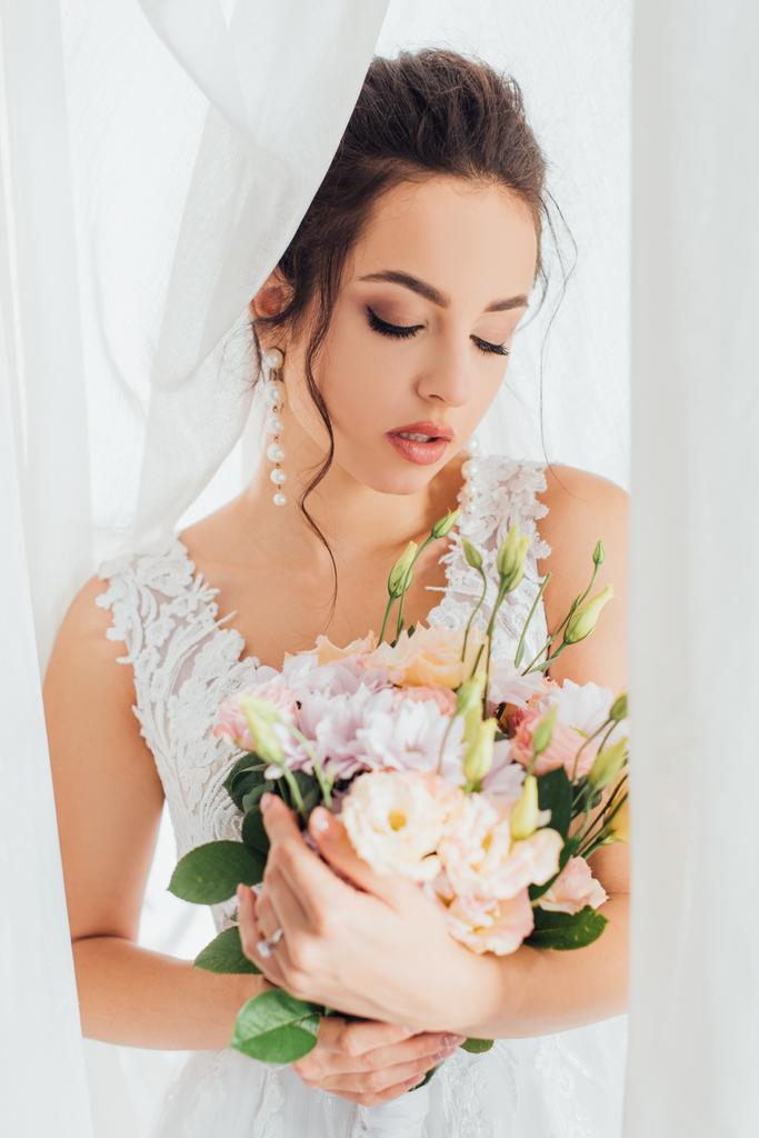 Foco seletivo da noiva no vestido de noiva segurando buquê perto de cortinas brancas
  - Foto, Imagem