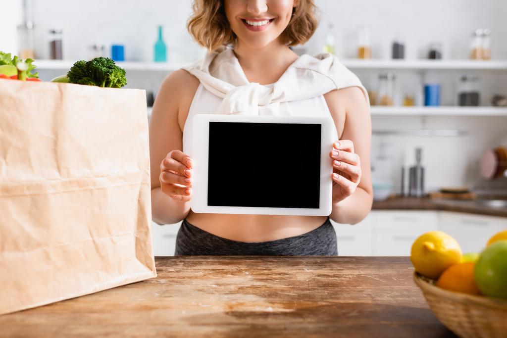 食料品と紙袋の近くに空白の画面を持つデジタルタブレットを保持する女性のクロップドビュー  - 写真・画像