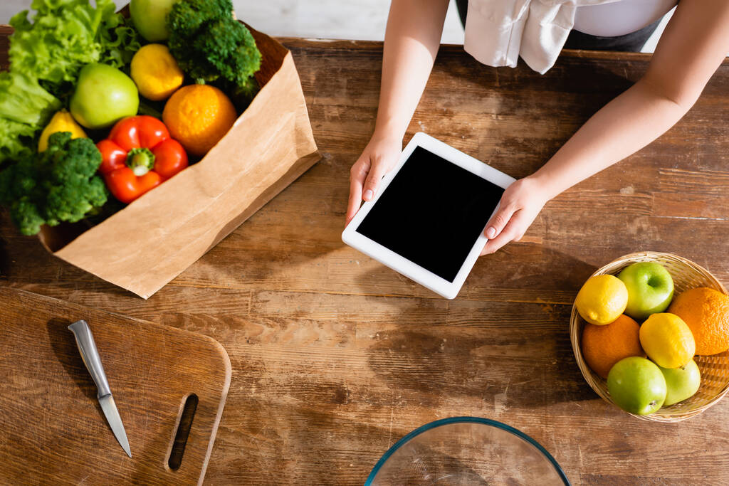 horní pohled ženy držící digitální tablet s prázdnou obrazovkou v blízkosti papírové tašky s potravinami a ovocem  - Fotografie, Obrázek