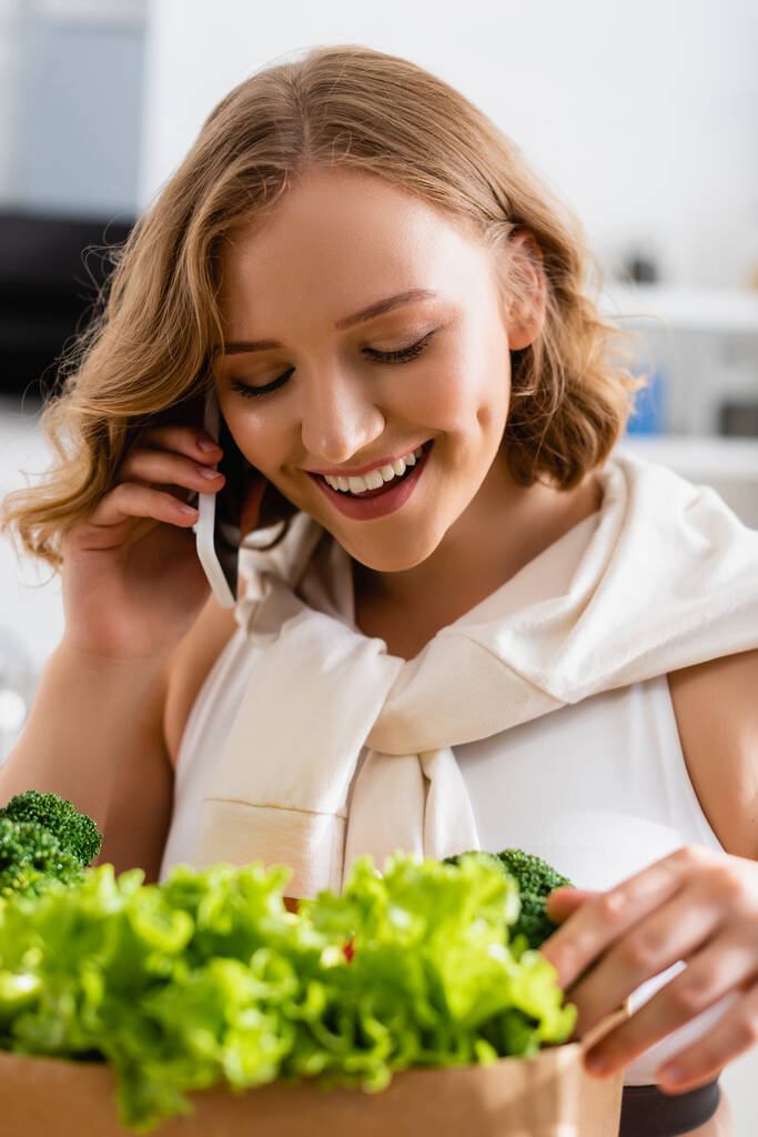 selective focus of young woman touching fresh lettuce near broccoli while talking on smartphone  - Photo, Image